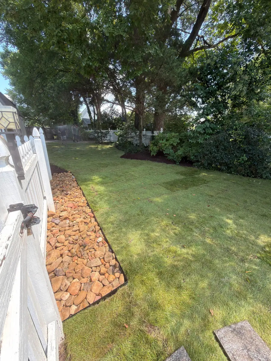 Sod installation in a fenced Concord, NC backyard corner with river rock edging and mature shade trees